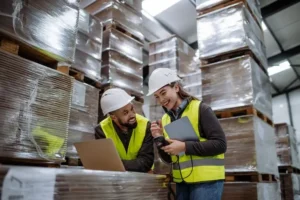Two construction procurement specialists in safety gear using a laptop and scanner to manage supply chain logistics and material inventory within a large warehouse.